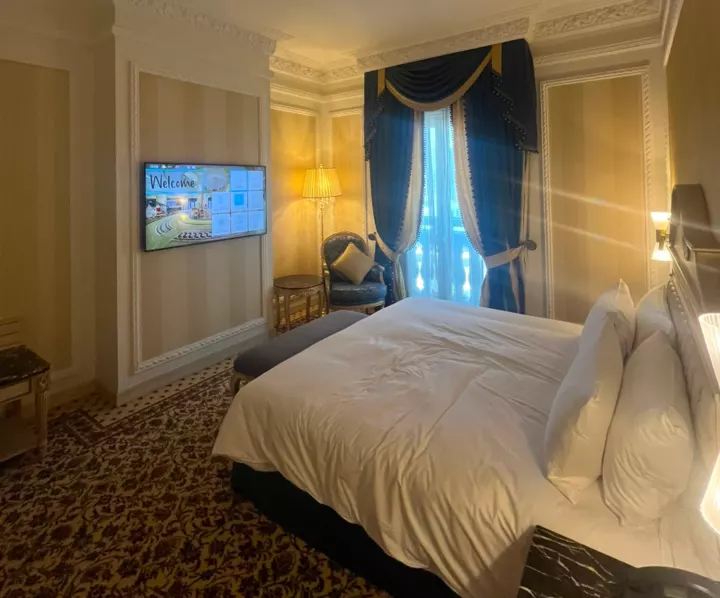 An ornate hotel bedroom with blue curtains, gold lamps and a large white bed.