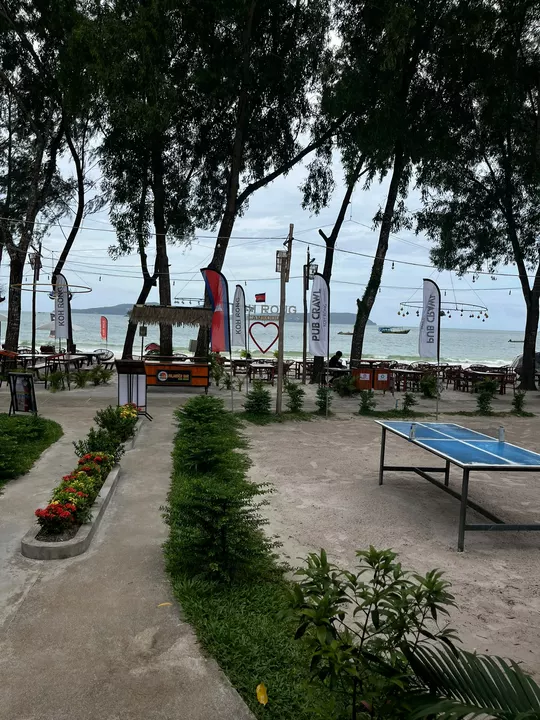 A sandy beachfront bar and garden with a table tennis table, trees and the sea beyond.
