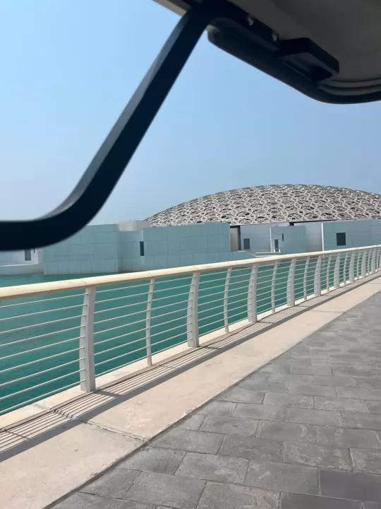 The silver lattice dome of Louvre Abu Dhabi beyond turquoise water and a sunlit walkway.