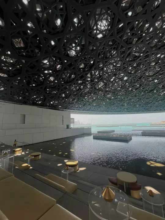 The patterned Louvre Abu Dhabi dome casting pools of sunlight over water and seating below.