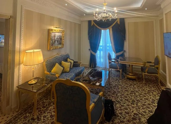 An ornate hotel sitting room with blue curtains, a chandelier, gold lamps and patterned carpets.