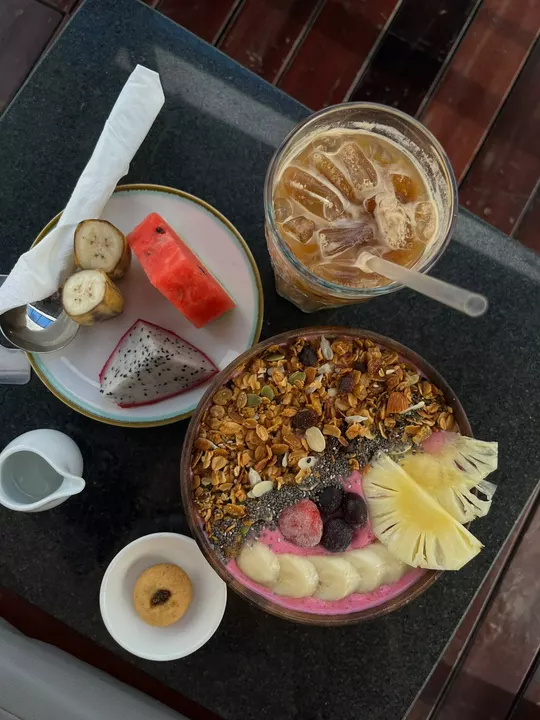 A colourful smoothie bowl topped with granola, pineapple, banana and berries, beside fruit and an iced coffee.