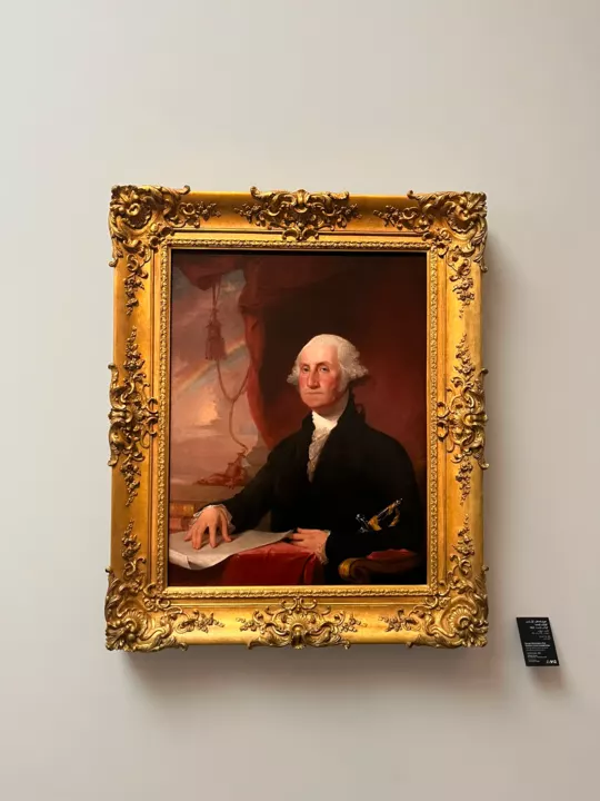 A portrait of George Washington in an ornate gold frame on a white museum wall.